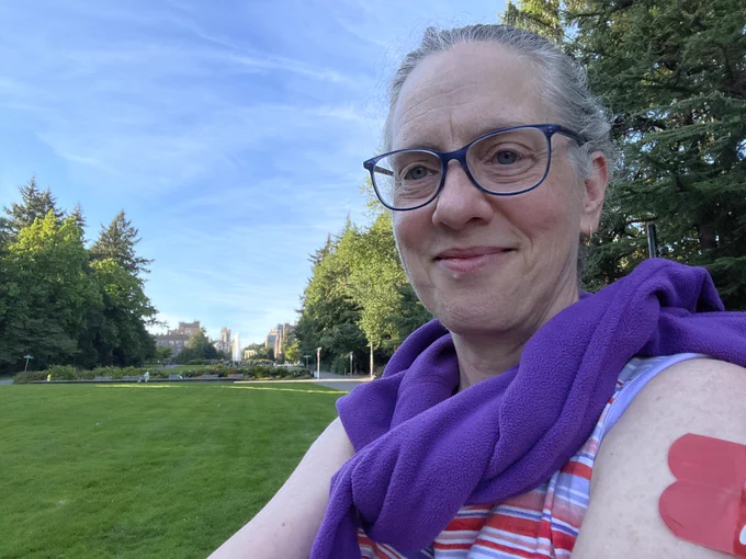 The photo shows a smiling professor on the lawn of Rainier Vista at the University of Washington in Seattle. The sun is shining, the sky is blue with wispy clouds, the trees and grass are bright green... it is a perfect day, really. The professor is wearing glasses, a sleeveless shirt, a purple sweater over her shoulders, and two red band-aids on her left arm, one where she got a covid vaccination booster and one where she got an influenza vaccine booster. In the background is a white column of water from Drumheller Fountain, and some buildings of upper campus.