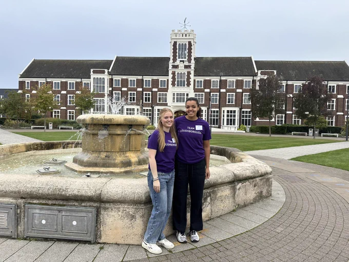 Two people in purple team Loughborough T-shirts smiling in front of the Hazlerigg building and fountain