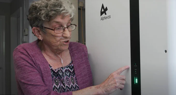 A photo of an older woman next to the battery in her home, that stores solar energy generated during the day.