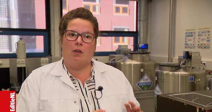 Een vrouw in een laboratoriumjas en bril die spreekt in een laboratorium, met wetenschappelijke apparatuur zichtbaar op de achtergrond.