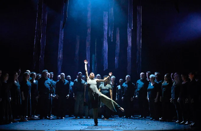 A scene from an opera. A large group of people in dark clothing stand in a semi circle on a dimly lit stage. In front of them two people are bathed in a spotlight. One wears a lighter dress and reaches up skywards.