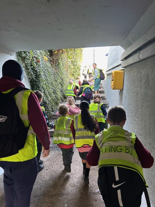 From behind, we see a group of schoolchildren in hi-vis jackets, and some teachers, walking through an underpass and up the steps into the daylight