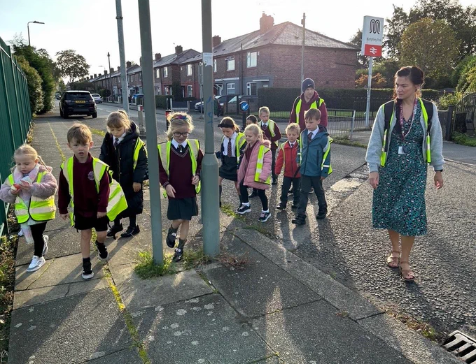 A group of primary school children in hi-vis jackets cross a road together on a sunny day, whilst a teacher walking beside them keeps watch
