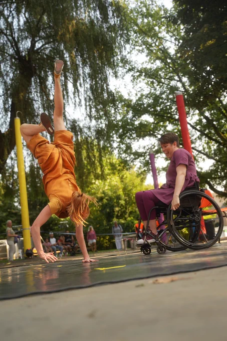 ALT Text: [main image]
An image of two acrobats captured during a performance of Look Mum, No Hands!  The first acrobat, Danielle, a white woman with ginger hair in ponytails, is wearing a yellow jumpsuit while she is performing a handstand on stage. While the second acrobat,  Kat, white woman with short brown hair and blue eyes a wheelchair user, dressed in a plum top and leggings, is photographed mid-movement as she movesa cross the stage. The duo are performign outdooes surronded by greenery. 