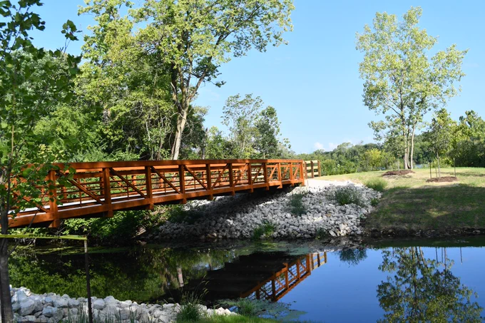 A rust-colored metal footbridge extending across a calm body of water. The water reflects the surrounding trees, the sky, and the bridge itself, creating a mirror-like effect.