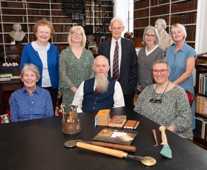 Eight people seated and standing behind a table, on which bookbinding tools and book bindings are displayed.
