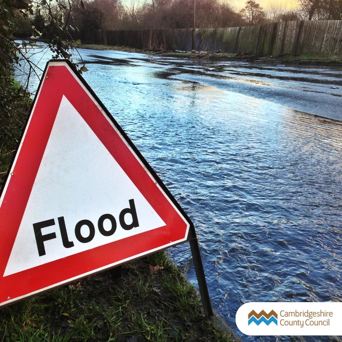 Images shows a flooded road with a red warning sign with 'flood' written on it.