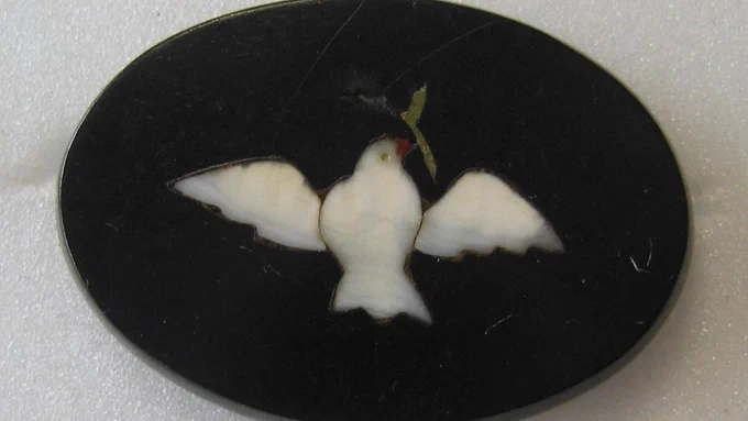 Colour photograph of a small Ashford Black Marble slip. It is oval in shape with a smooth, shiny black surface. The decoration is minimal and consists of one white dove with wings outspread, carrying an olive branch. His beak is a deep orange colour and the branch is a beautiful shade of green.