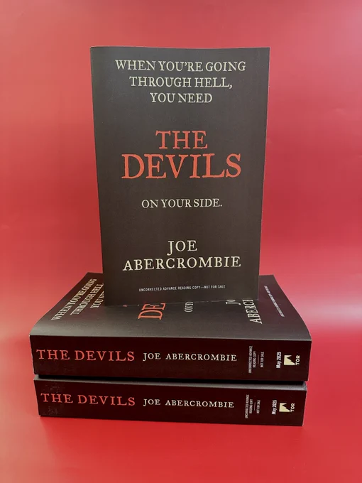 Three ARCs of The Devils by Joe Abercrombie are stacked in front of a red background