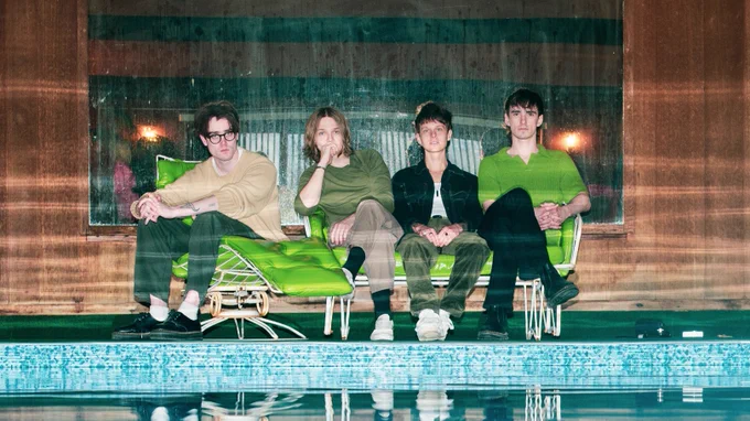 An image of the members of Hippo Campus, wearing various beige, green, and black outfits, sitting on green lounge chairs in front of a brown wood background near the edge of a pool.