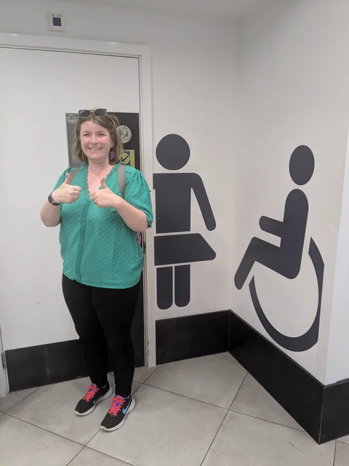 Clare stands outside a Changing Places toilet in Horsham Swan Walk Shopping Centre. She is smiling at the camera and giving two thumbs up.