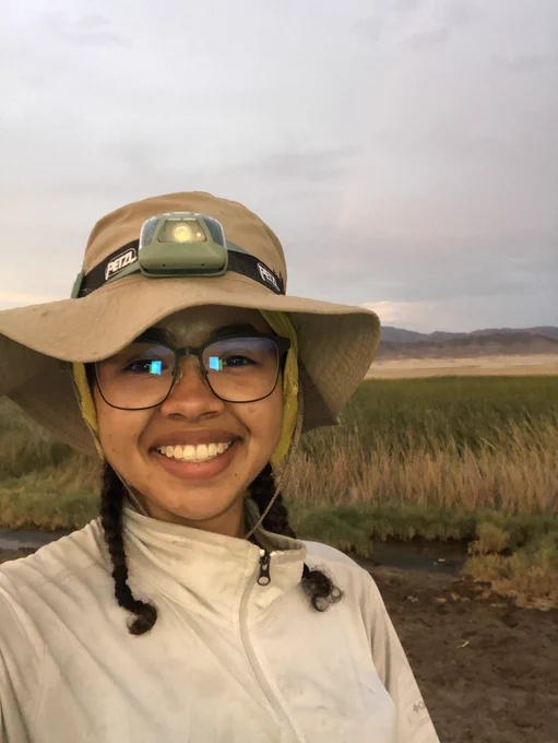 Me, a young Black woman smiling so hard. I’m wearing my headlamp and sun hat, my white zip up is pretty dirty from being in the field. The background features a lush desert wetland and mountains.
