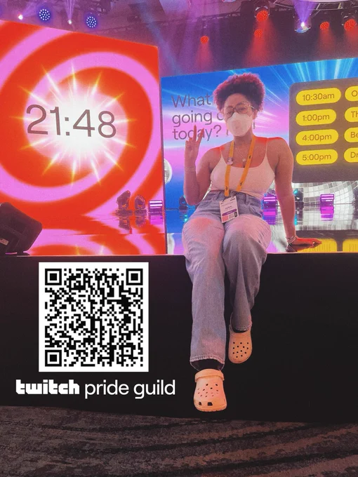 Sage sitting on the main stage with jeans, crocs and a white crop top on. on the bottom left of the image is a QR code with the Twitch Pride Guild logo underneath.
