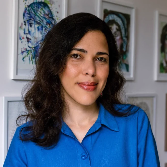A photograph of Nicole Louie wearing a blue shirt and standing in front of a series of white frames on a white wall. 
