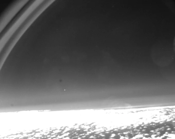 A black-and-white image showing Earth's surface at the bottom and a bright dot in the distance above Earth's surface. At the top left of the image are three curved lines.