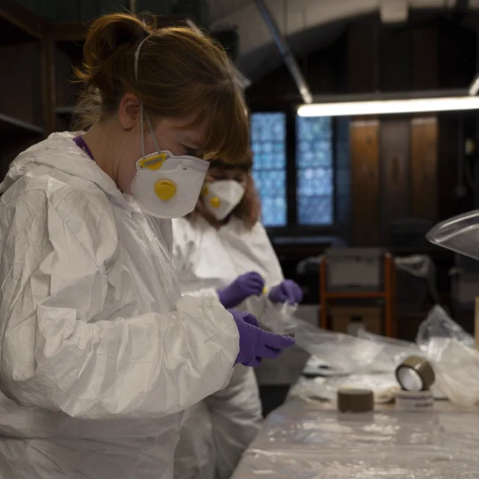 Archivist Jessica Smith and Conservator Laura Snow dressed in white hazmat suits and face masks prepping reel to reel tapes.