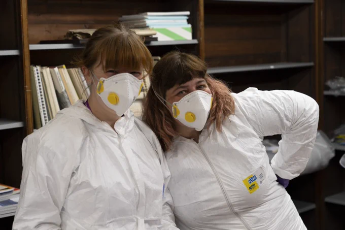 Curator Jessica Smith and Conservator Laura Snow dressed in face masks and white hazmat suits smiling at camera.