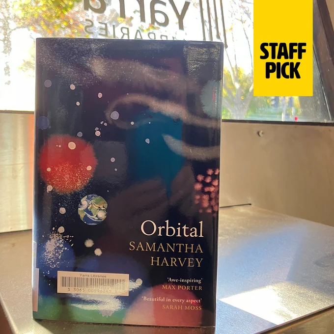 Staff pick book 'Orbital' by Samantha Harvey displayed in a library window, with sunlight filtering through trees in the background.