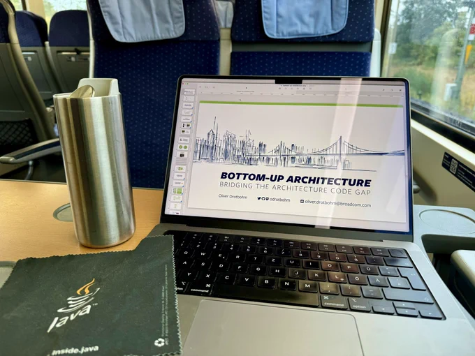 A laptop standing open on a train desk showing a presentation on software architecture. A coffee coffee mug and a microfiber towel sporting a Java (programming language) logo right next to it.