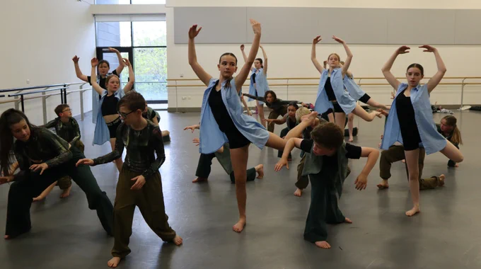 Dancers perform in grey studio, six dancers in blue waistcoats and black garments raise their arms in the air. Other dancers in the image are seen in kneeling dance position wearing dark green tops and dark green trousers. 