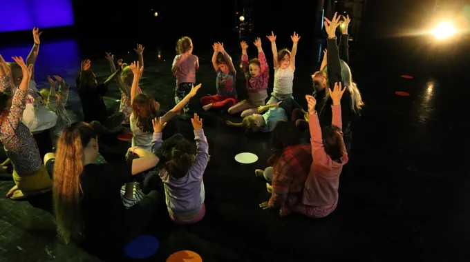 young dancers sat on stage floor with arms raised.