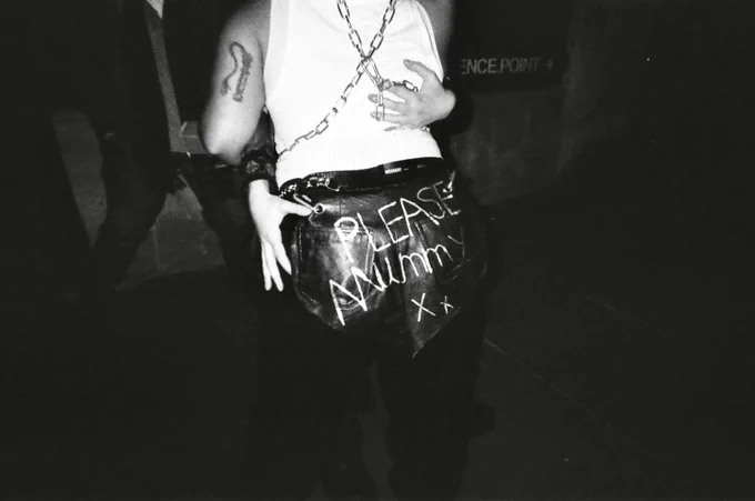 Black and white photo from a party. Two people are hugging, we only see the back of one of them. They’re wearing black leather trousers with the text ‘please mummy xx’, a white tank top and chains