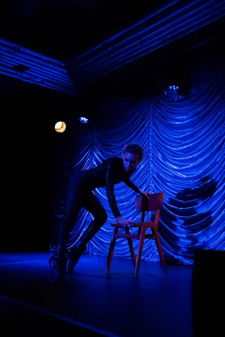 Photo from a performance. A performer with short hair wearing tight black clothes and stiletto heels is on stage and using a chair as a prop