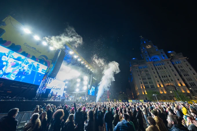 A live, outdoor concert at night with a large crowd. The Royal Liver Building can be seen in the background.