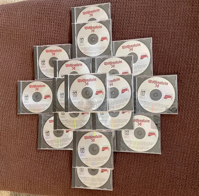 A collection of Wolfenstein 3D CDs arranged in a cross-like pattern on a textured brown surface. Each CD is in its original packaging, featuring the Wolfenstein 3D logo in red, the id Software and Apogee logos, and text detailing the game’s content. The reflective surfaces of the CDs add a slight shine to the image, giving it a nostalgic, retro-gaming vibe.