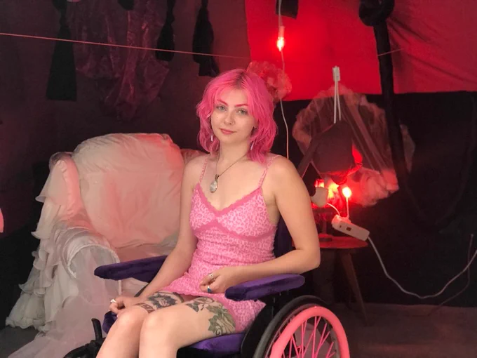 Image of a young woman in a wheelchair with bright pink hair wearing a bright pink dress.
