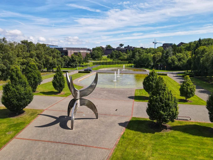 A photograph of a steel artwork on a large paved plaza surrounded by grassy lawns. The sculpture is comprised of three curved legs on the bottom, which are mirrored on the top. There is also a large pond with fountains in it