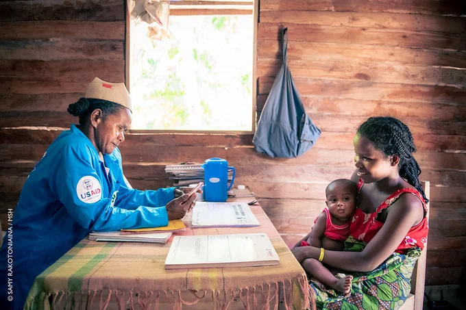 Two women sit across each other at a table. One woman is holding a small child in her arms. 