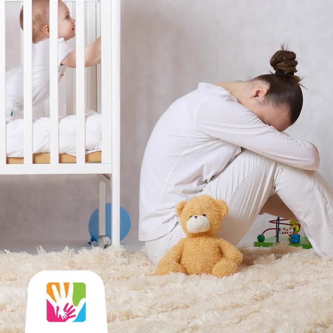 An image of a woman with her head in her lap while her baby is in a white crite trying to get her attention.