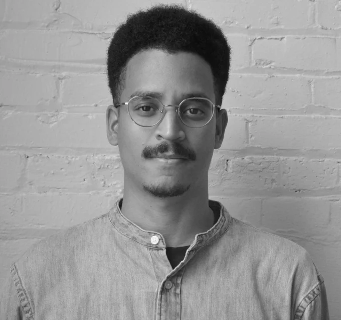 A greyscale profile photo of Tura Cousins Wilson against a whitewashed brick background. His gaze is both direct and relaxed.