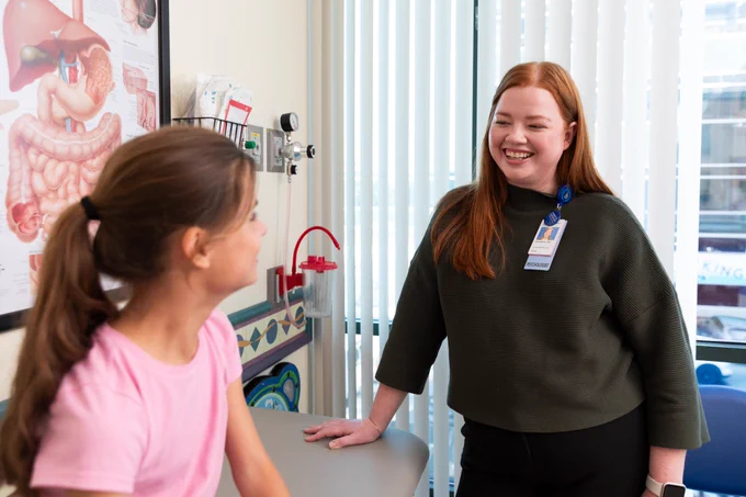 Dr. Dana Bakula stands next to an exam table. Her hands rests on the table. Sitting on the table is a young female patient. They look at each other and smile. 
