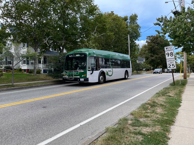 A gray and forest green battery electric bus identified as "Transit" with an image of an AC power cable and plug running across the side. On the side of the road, signs read "WEST 98" and "SPEED LIMIT 25".