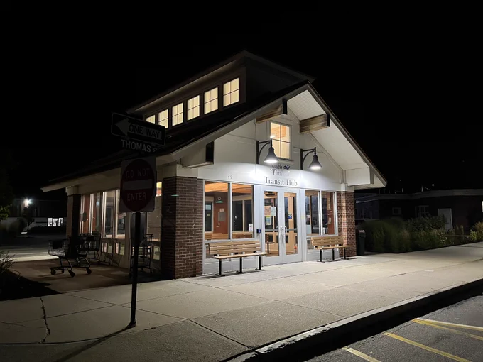 Building in a similar form to a single-story, single-family home with a gabled roof/attic reads "South Portland Transit Hub". The building has large glass windows on all sides and is well-lit in the night.