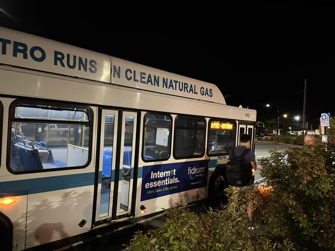 At night, two passengers prepare to board a white-painted bus reading "METRO RUNS ON CLEAN NATURAL GAS" in teal along the top. The digital destination sign reads "1 PTC - AMTRAK".