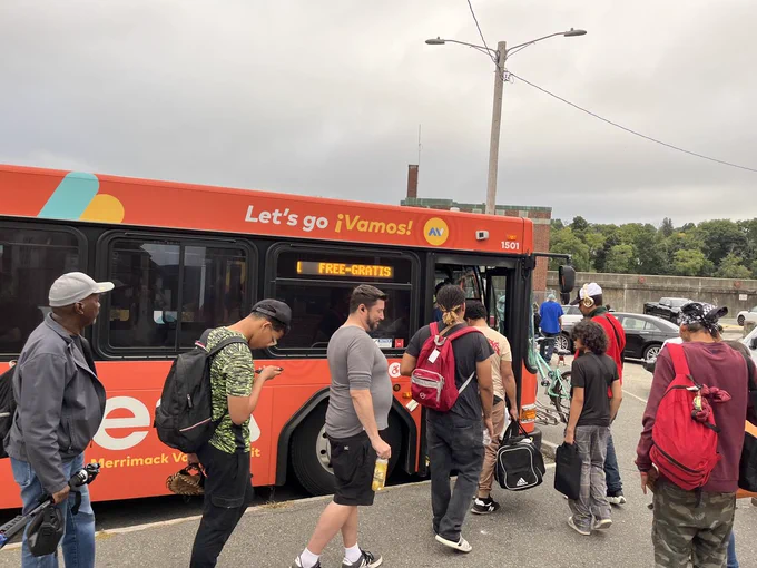 Approximately 10 people line up to board an orange bus operated by MeVa, Merrimack Valley Transit. The bus has "Let's go ¡Vamos!" written on it, and its digital destination sign reads "1 FREE-GRATIS".