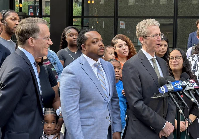 Marcel Brown, a 34-year-old black man in a blue suit, stands at a press conference surrounded by attorneys and supporters. 