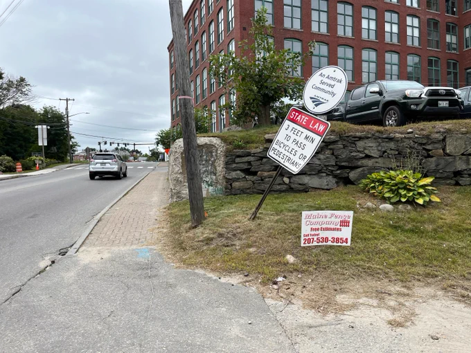 A road appears with a somewhat narrow sidewalk at its right side. To the right of the sidewalk and near a telephone pole, a sign post appears to be bent about 30 degrees. On the pole are mounted two signs: The top one reads "An Amtrak Served Community" with the Amtrak logo. The bottom sign reads "STATE LAW - 3 FEET MIN TO PASS BICYCLES OR PEDESTRIANS".