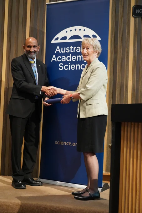 A person receiving a certificate in a cylinder from another person. They stand shaking hands in front of a banner with the logo of the Australian Academy of Science. 