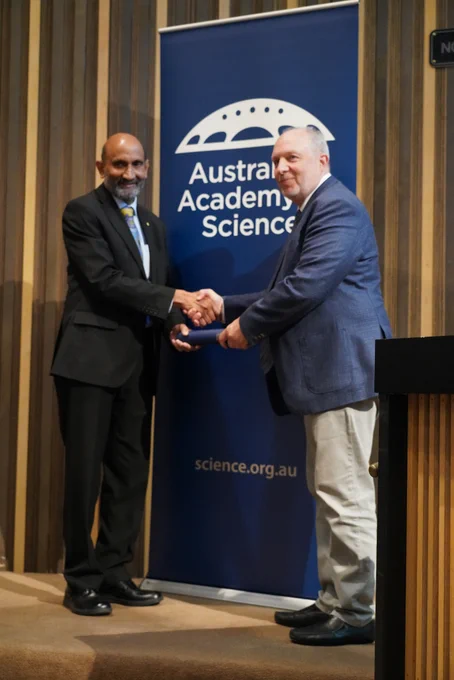 A person receiving a certificate in a cylinder from another person. They stand shaking hands in front of a banner with the logo of the Australian Academy of Science. 
