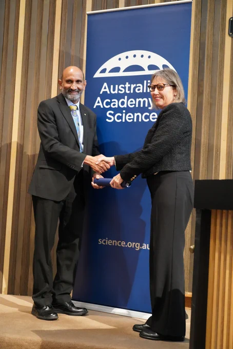 A person receiving a certificate in a cylinder from another person. They stand shaking hands in front of a banner with the logo of the Australian Academy of Science. 