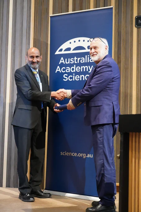 A person receiving a certificate in a cylinder from another person. They stand shaking hands in front of a banner with the logo of the Australian Academy of Science. 