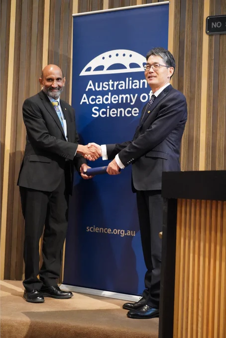 A person receiving a certificate in a cylinder from another person. They stand shaking hands in front of a banner with the logo of the Australian Academy of Science. 