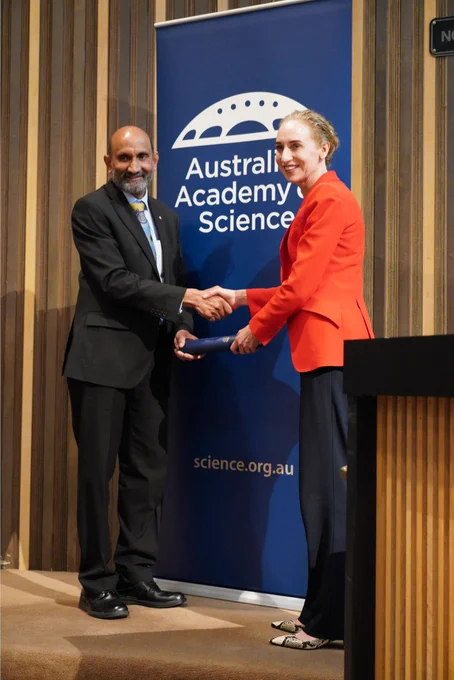 A person receiving a certificate in a cylinder from another person. They stand shaking hands in front of a banner with the logo of the Australian Academy of Science. 