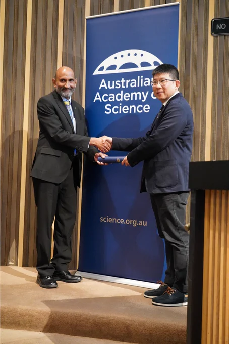 A person receiving a certificate in a cylinder from another person. They stand shaking hands in front of a banner with the logo of the Australian Academy of Science. 