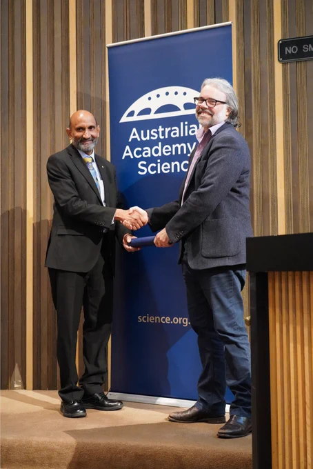 A person receiving a certificate in a cylinder from another person. They stand shaking hands in front of a banner with the logo of the Australian Academy of Science. 