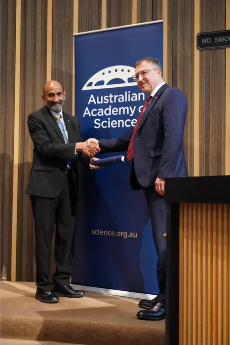 A person receiving a certificate in a cylinder from another person. They stand shaking hands in front of a banner with the logo of the Australian Academy of Science. 